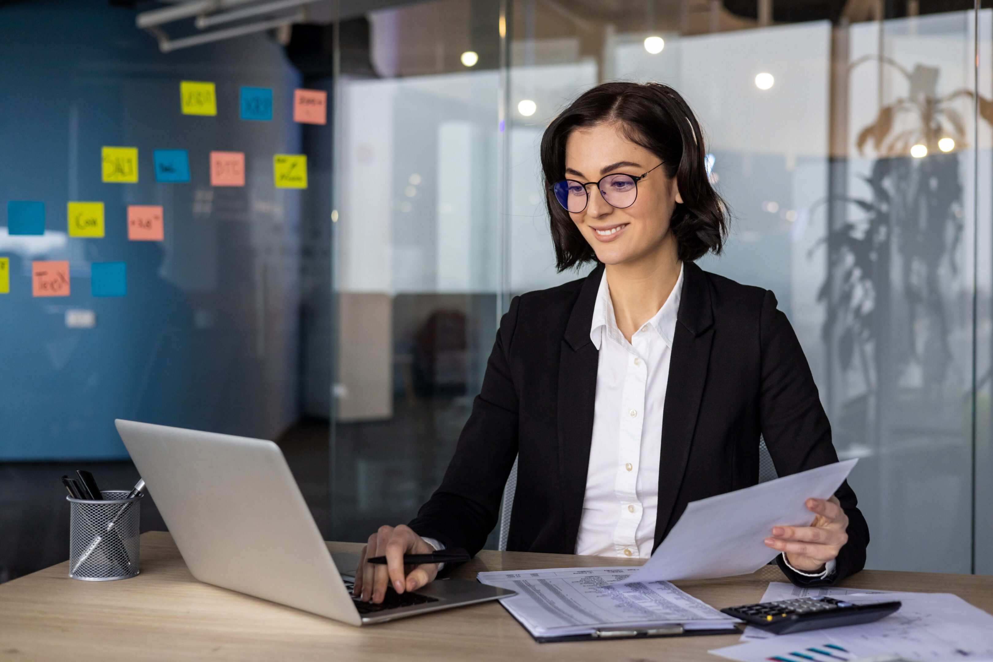 Woman in business office wearing glasses and headphones, working on laptop with documents. Shows productivity, focus, and engagement in professional tasks amid modern office setting.